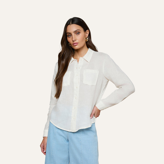 A woman wearing the Isla Linen Shirt—a white linen button-down with two front pockets—paired with light blue pants, stands against a plain light background with her hand on her hip.
