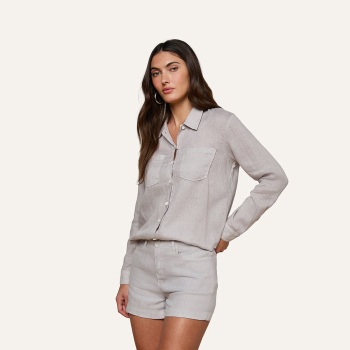 A woman with long brown hair wears the Isla Linen Shirt, a light beige button-down style with front pockets, paired with matching shorts. She stands against a plain, light background, looking toward the camera in a relaxed pose.