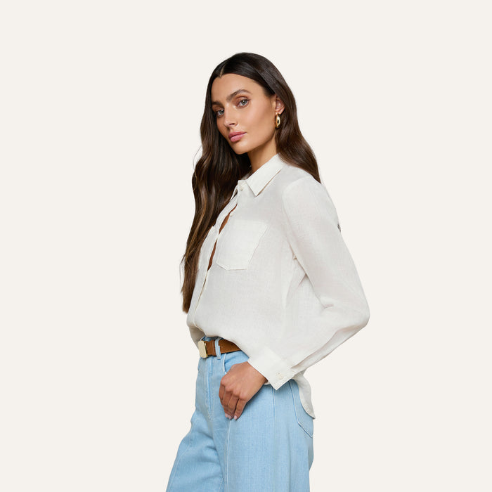 A woman with long brown hair, dressed in the Isla Linen Shirt—a white button-down style featuring chest pockets—paired with light blue jeans, stands against a plain light background, looking confidently at the camera.