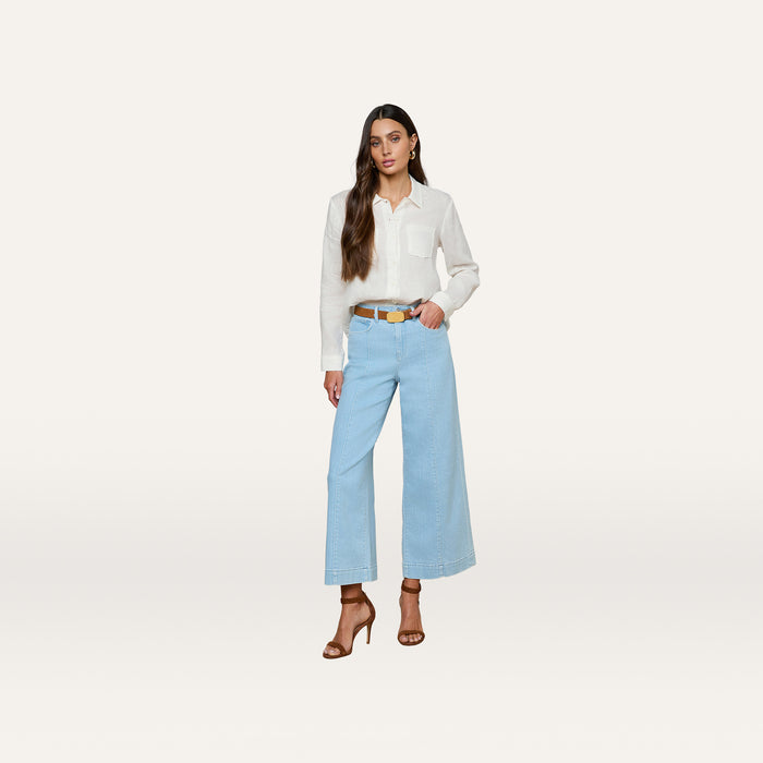 A woman stands against a plain white background, wearing the Isla Linen Shirt with long sleeves, paired with light blue wide-leg jeans, a brown belt, and brown high-heeled sandals. She has long brown hair and a neutral expression.