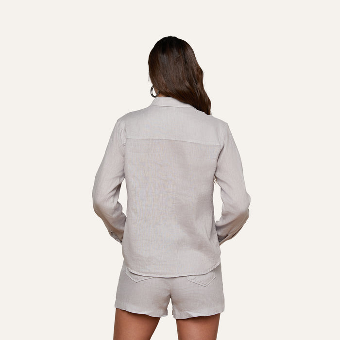 Wearing the Isla Linen Shirt, a woman with long brown hair is seen from behind in a light-colored ensemble, standing against a plain light background.