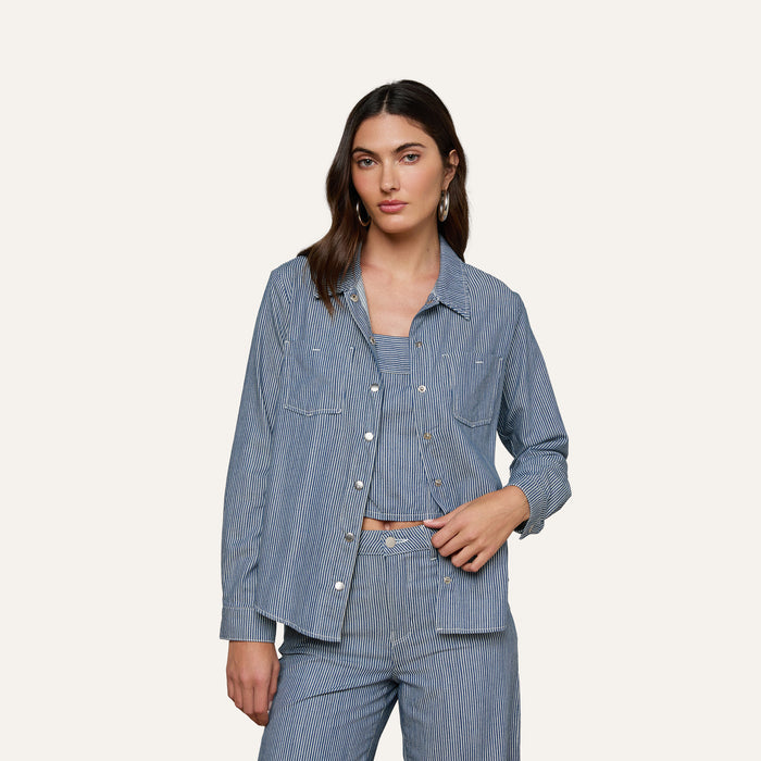 A woman wearing the Isla Shirt by L’AGENCE in featherweight indigo pinstripe denim over a coordinating top and pants stands against a plain light background, looking at the camera with a relaxed expression.