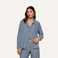 A woman wearing the Isla Shirt by L’AGENCE in featherweight indigo pinstripe denim over a coordinating top and pants stands against a plain light background, looking at the camera with a relaxed expression.