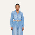 A woman stands facing forward against a plain off-white background, wearing the Isla Shirt styled as a lightweight, vintage-wash denim jumpsuit with a beige belt, hoop earrings, and her hair pulled back.