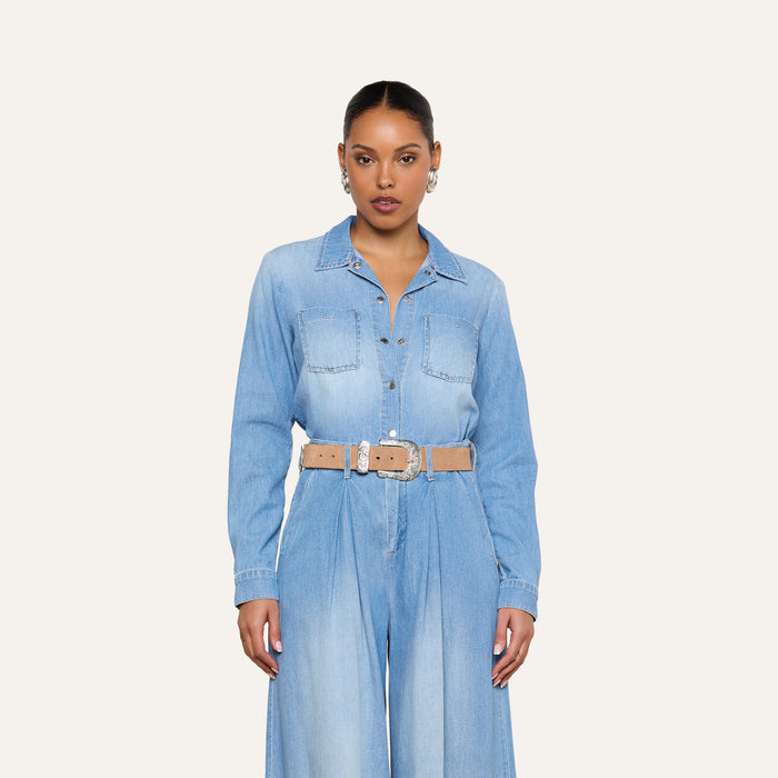 A woman stands facing forward against a plain off-white background, wearing the Isla Shirt styled as a lightweight, vintage-wash denim jumpsuit with a beige belt, hoop earrings, and her hair pulled back.