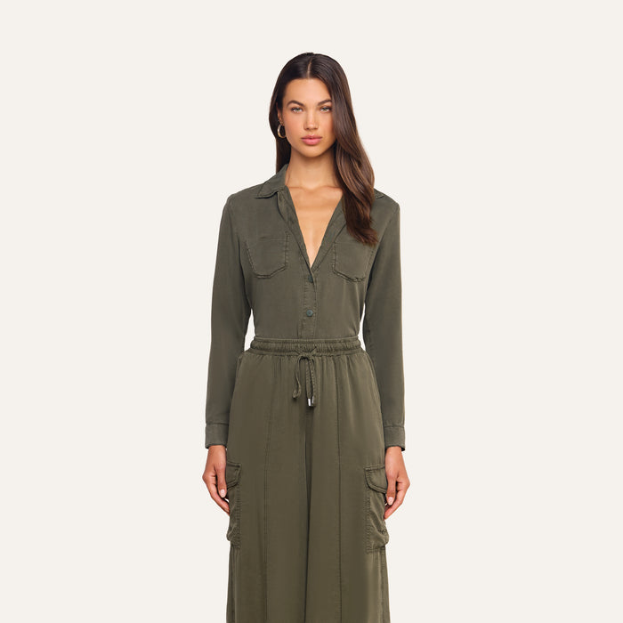 A woman stands facing forward, wearing the Isla Shirt in a long-sleeve olive green featherweight twill, featuring button details, a drawstring waist, and large front pockets. She has long brown hair and a neutral expression. The background is plain off-white.
