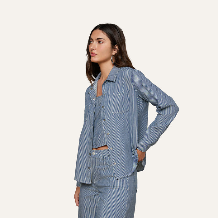 A woman stands against a plain background wearing the Isla Shirt in indigo pinstripe with matching pants, her left hand in her pocket and a neutral expression.