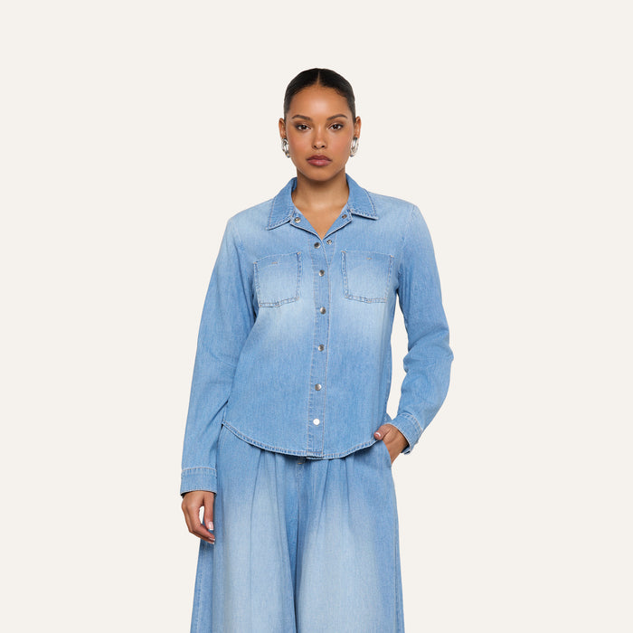 A woman wearing the Isla Shirt—a lightweight light blue denim shirt with front pockets—paired with matching wide-leg denim pants, stands against a plain off-white background.