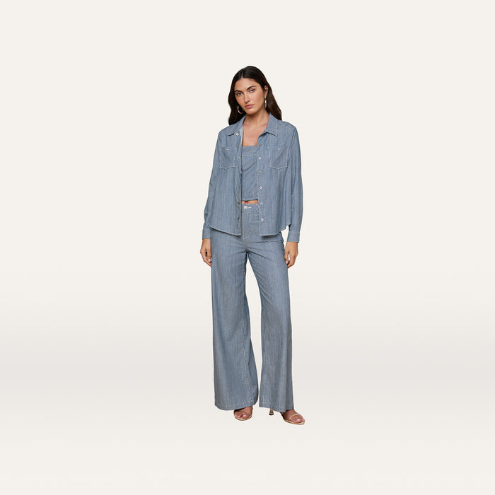 A woman stands against a plain white background wearing an indigo pinstripe pantsuit with wide-leg trousers, the Isla Shirt layered over a top, and open-toe heels.
