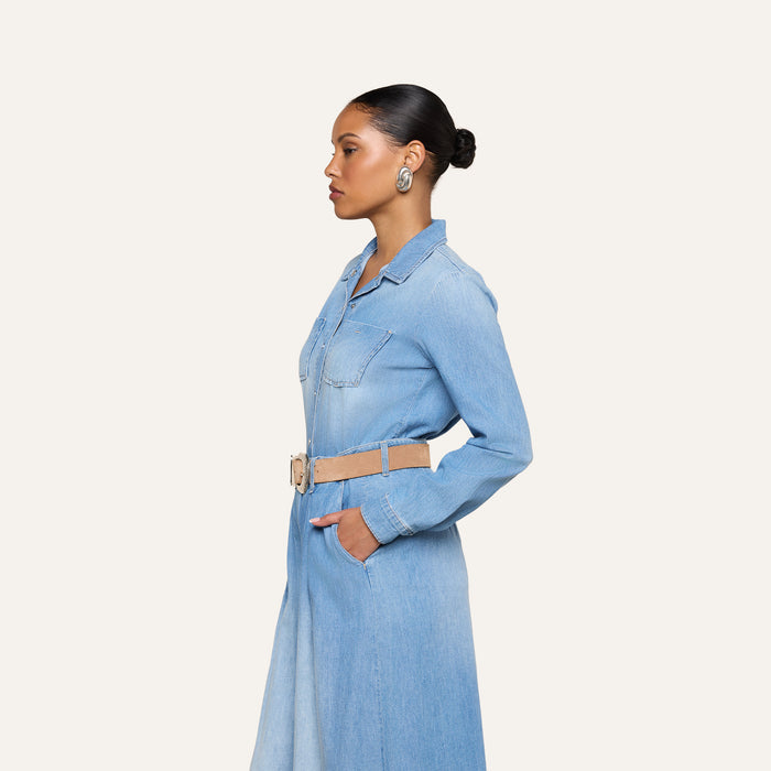 Woman in the Isla Shirt, a lightweight light blue vintage wash denim shirt dress with pockets, styled with a beige belt and large round earrings, stands in profile against a plain white background.