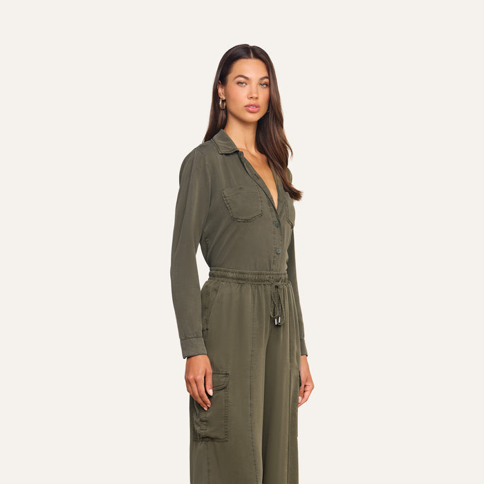 A woman wearing the Isla Shirt in olive green featherweight twill, featuring long sleeves, large front pockets, a collared neckline, and a drawstring waist, standing against a plain light background.