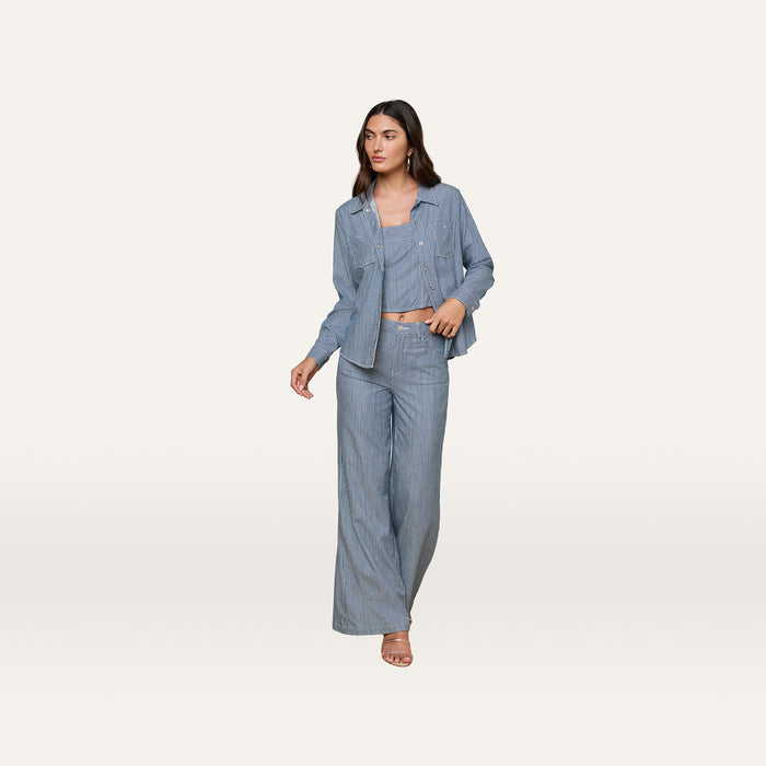 A woman stands against a plain, light background wearing the Isla Shirt in featherweight light blue denim, paired with a matching crop top and wide-leg pants. She looks off to the side.