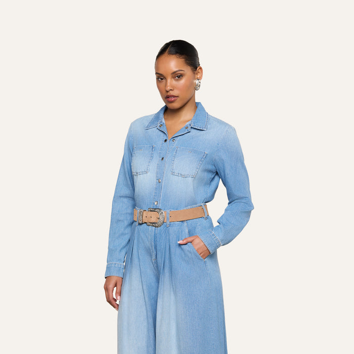 A woman stands against a plain white background, wearing the Isla Shirt—a lightweight, light blue denim jumpsuit with chest pockets and a tan belt—with one hand casually placed in her pocket.