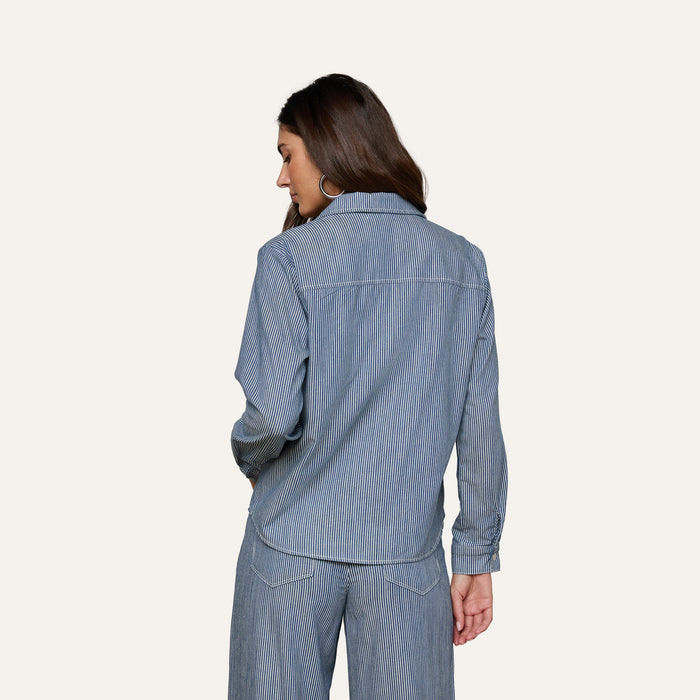 Woman standing with her back to the camera, wearing the Isla Shirt in indigo pinstripe featherweight denim from L’AGENCE, paired with matching pants, against a plain light background.