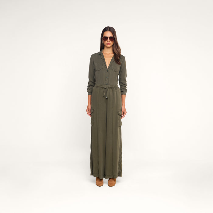 A woman wearing sunglasses and brown shoes stands facing forward against a plain white background, hands at her sides, modeling the Isla Shirt—a long-sleeve, olive green maxi dress made from featherweight twill with pockets.