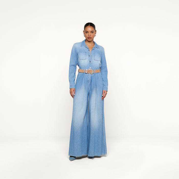 A person stands against a plain white background wearing the Isla Shirt—a lightweight light blue denim top styled with wide-leg jeans, a tan belt, and vintage-wash blue shoes.