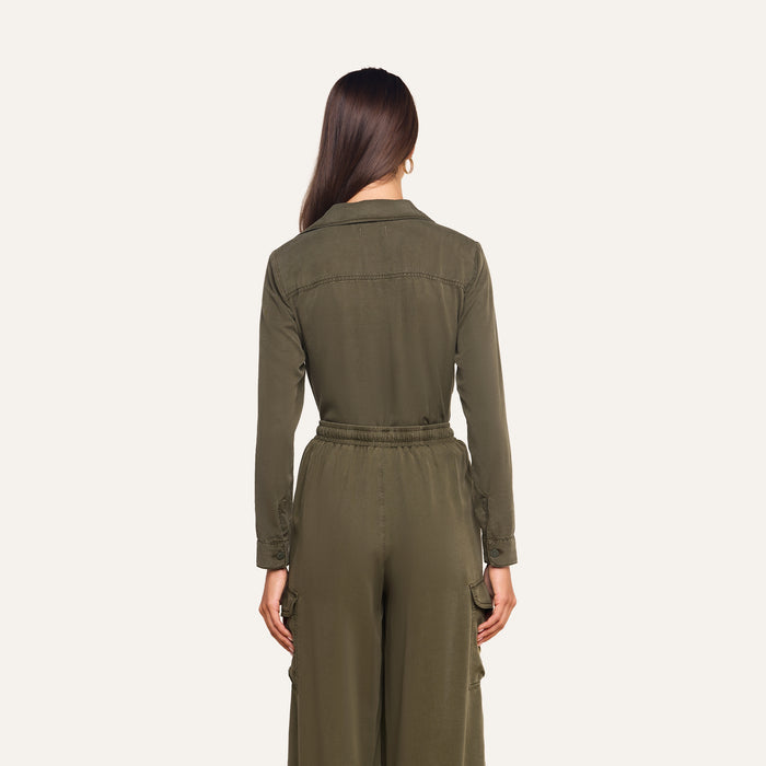 A woman with long brown hair stands facing away, wearing the Isla Shirt in olive green featherweight twill, featuring an elastic waistband and cargo pockets, against a plain light background.