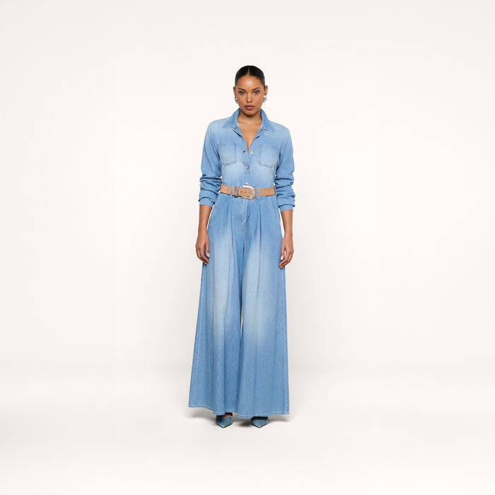 A woman stands against a plain white background wearing the Isla Shirt, styled with wide-leg light blue denim bottoms, a tan belt, and black pointed shoes.