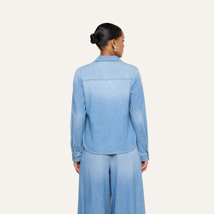 A woman with her hair in a bun, wearing the lightweight light blue Isla Shirt and matching wide-leg pants, is shown from the back against a plain, light background.