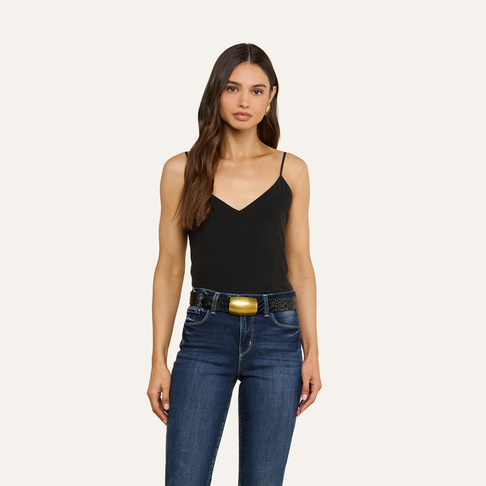 A woman with long brown hair wears the Jane Silk Camisole in black, featuring adjustable straps, paired with dark blue jeans and a wide black belt with a large gold buckle, standing against a plain light background.