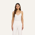 A woman with long brown hair wears the Jane Silk Camisole, a sleeveless white top with adjustable straps, paired with high-waisted white pants. She stands against a plain light background, facing forward with a neutral expression.