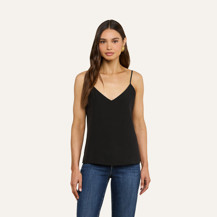 A woman with long brown hair wears the Jane Silk Camisole—a black, bias-cut, silk V-neck top—paired with blue jeans, standing against a plain light background.