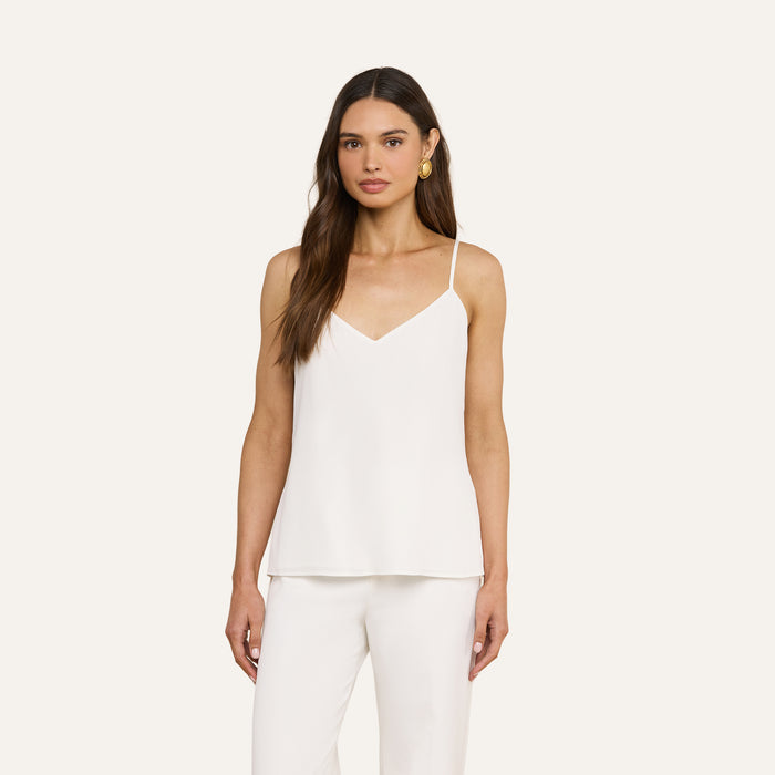 A woman with long brown hair wears the Jane Silk Camisole, featuring adjustable straps, paired with matching white pants. She faces forward against a plain light background and accessorizes with gold hoop earrings.