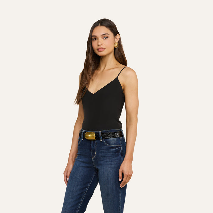 A woman wearing the Jane Silk Camisole with adjustable straps and blue jeans stands against a plain light background. She has long brown hair, gold hoop earrings, and a black belt with a large gold buckle.
