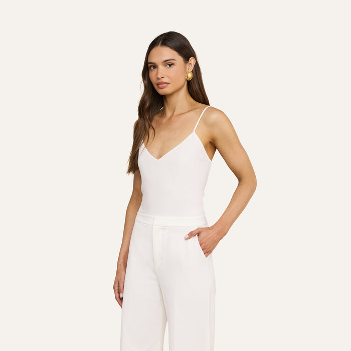 A woman with long brown hair wears the Jane Silk Camisole, a sleeveless white top with adjustable straps, paired with white pants as she stands with one hand in her pocket against a plain light background.