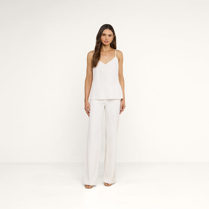 A woman with long brown hair wears the Jane Silk Camisole—a bias-cut silk top with adjustable straps—paired with white wide-leg pants, standing against a plain white background with a neutral expression.