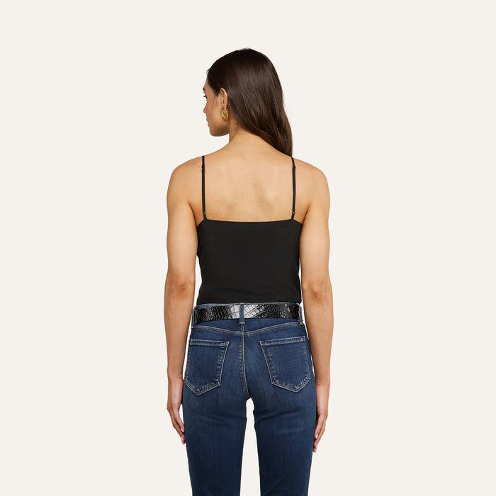 A woman with long brown hair stands facing away, wearing the Jane Silk Camisole with adjustable straps, paired with dark blue jeans, a black belt, and gold hoop earrings against a plain light background.