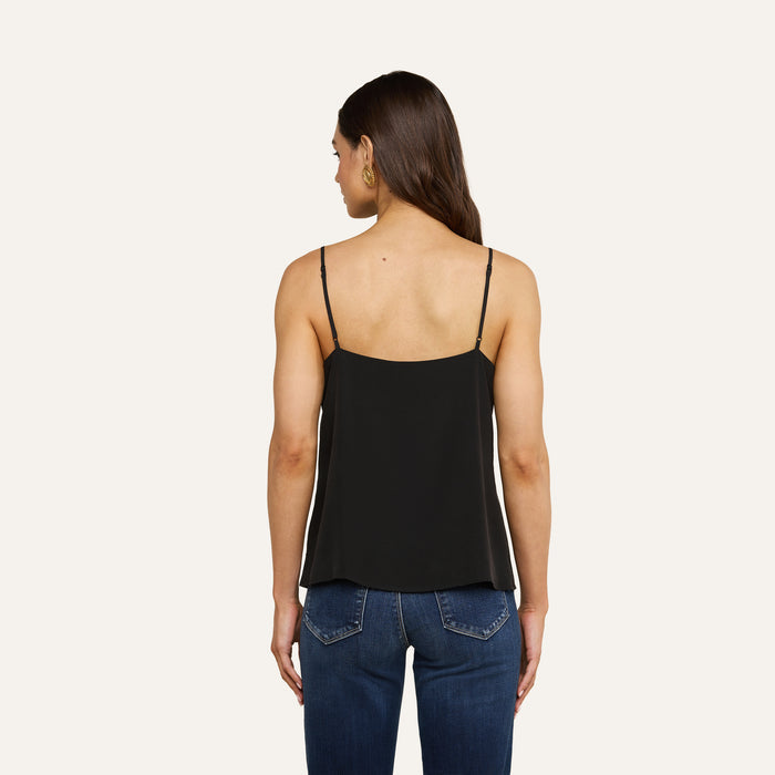 A woman stands with her back to the camera, wearing the Jane Silk Camisole in black—featuring adjustable straps—paired with blue jeans. She has long dark hair worn down and gold hoop earrings, set against a plain, light-colored background.