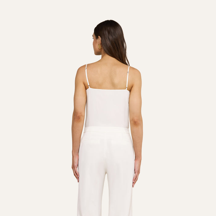 A woman with long brown hair stands facing away against a plain light background, wearing the Jane Silk Camisole—a bias-cut silk top with adjustable straps—paired with white pants.