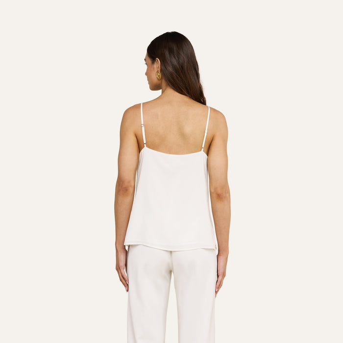 A woman with long brown hair stands facing away, wearing the Jane Silk Camisole with adjustable straps and matching white pants, set against a plain light background.