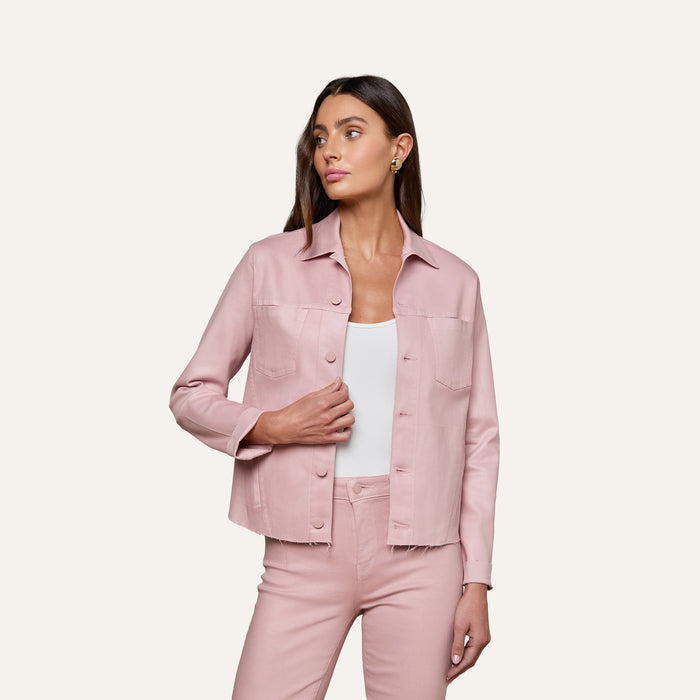 A woman wearing the Janelle Coated Jean Jacket in light pink with matching slim fit pants over a white top stands against a plain light background, looking to her left with one hand by her side and the other on her hip.