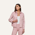 A woman wearing the Janelle Coated Jean Jacket in light pink with matching slim fit pants over a white top stands against a plain light background, looking to her left with one hand by her side and the other on her hip.