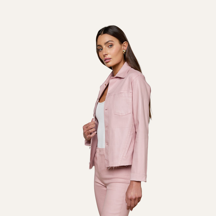 A woman with long brown hair wears the Janelle Coated Jean Jacket in light pink over a white top and matching pink pants, standing against a plain off-white background. She faces slightly to the side, looking forward with a neutral expression.