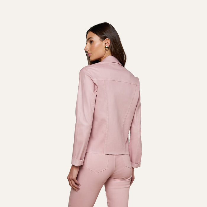 A woman with long brown hair wears the slim fit Janelle Coated Jean Jacket in light pink with matching pants, standing against a plain off-white background and looking to her left.