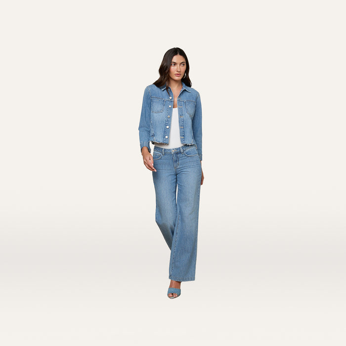 A woman stands against a light background, wearing the slim fit Janelle Jean Jacket, a white top, blue jeans, and blue heeled sandals.