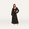 A woman stands barefoot against a plain white background, wearing the Janus Cover-Up Robe—a long-sleeve, sheer black lace bohemian layer—over a black bikini.