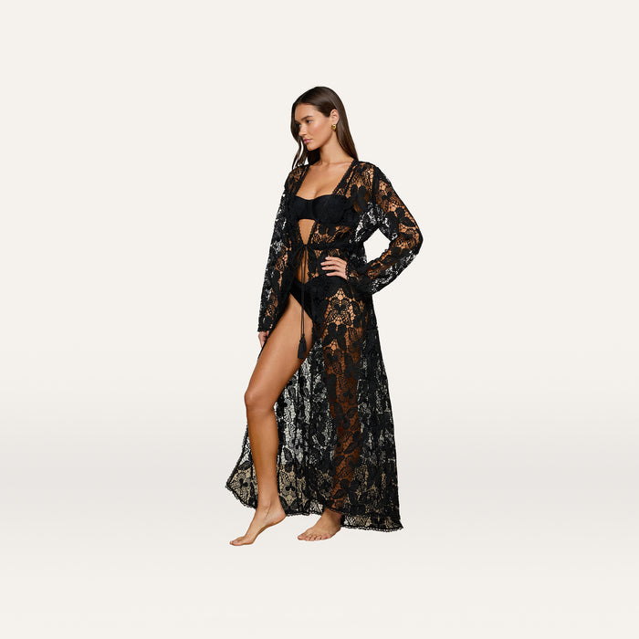 A woman stands barefoot wearing the Janus Cover-Up Robe—a long, sheer black lace piece reminiscent of a bohemian cover-up—styled over black lingerie and posing against a plain, light background.