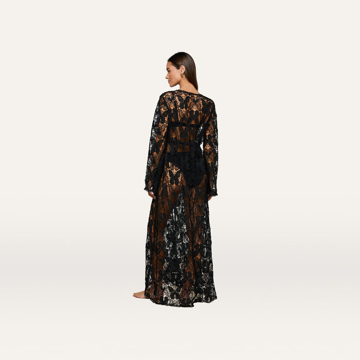 A woman stands barefoot, facing away, wearing the Janus Cover-Up Robe—a long, sheer black lace maxi beach dress with long sleeves. The robe features a floral pattern and reveals her silhouette against a plain white background.