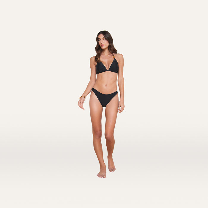 A woman stands barefoot against a plain white background, wearing a black bikini with a triangle top and the Jean Bikini Bottom, which features a high-leg cut and provides full coverage. She has long brown hair and a neutral expression.