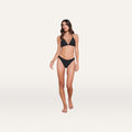 A woman stands barefoot against a plain white background, wearing a black bikini with a triangle top and the Jean Bikini Bottom, which features a high-leg cut and provides full coverage. She has long brown hair and a neutral expression.