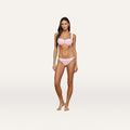 A woman stands against a plain white background, wearing the Jean Bikini Bottom—a high-leg cut bikini bottom crafted from textured bouclé fabric in light pink with white straps and gold accent details—looking at the camera with a neutral expression.