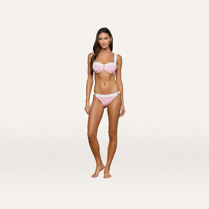 A woman stands against a plain white background, wearing the Jean Bikini Bottom—a high-leg cut bikini bottom crafted from textured bouclé fabric in light pink with white straps and gold accent details—looking at the camera with a neutral expression.