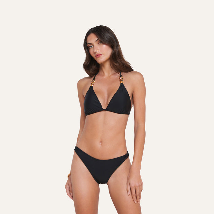 A woman models the Jean Bikini Bottom, featuring a high-leg cut and gold chain details on the black straps, against a simple light backdrop.
