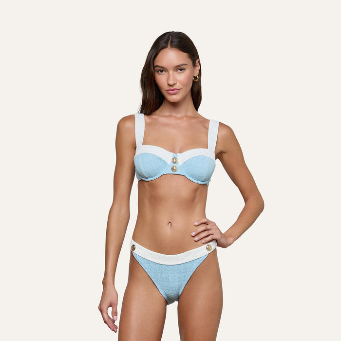 Woman standing against an off-white background, wearing the Jean Bikini Bottom—a light blue textured bouclé swim bikini with wide white straps and gold button accents, featuring a high-leg cut. One hand rests on her hip.