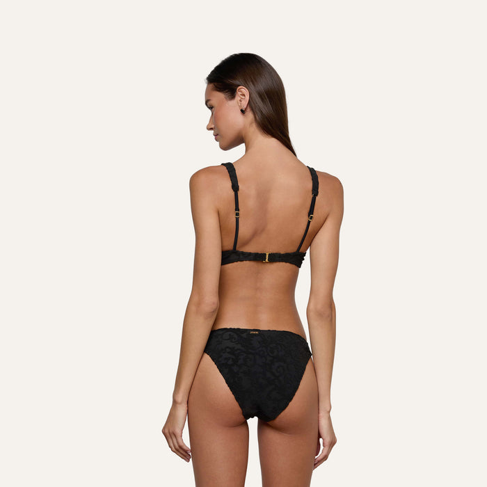 A woman with long brown hair stands with her back to the camera, wearing a timeless black lace bra and the Jean Bikini Bottom, set against a plain light background.