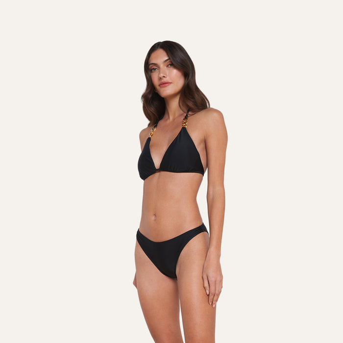 A woman stands against a plain background, wearing the Jean Bikini Bottom paired with a black bikini featuring chain-detail straps and a high-leg cut, gazing slightly to the side with a relaxed expression.